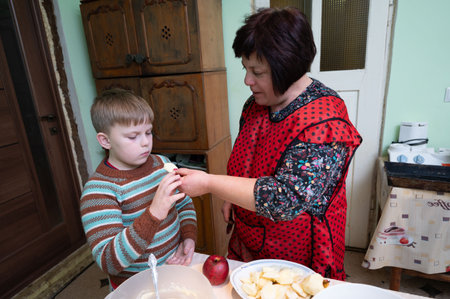 A woman helps her young son slice apples in a warm kitchen adorned with vintage decor, both immersed in the joy of cooking and creating memories on a peaceful day.の写真素材