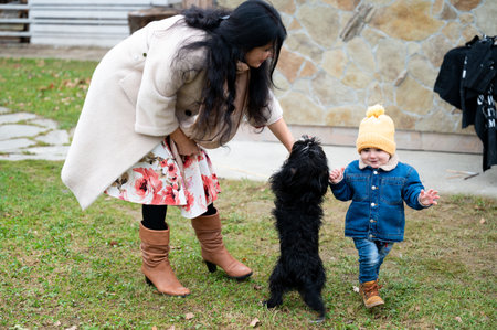 A woman pets a black Griffon Bruxellois puppy while a toddler in a yellow hat stands beside, seemingly taking steps with the assistance of the dog's joyful presence on grass.の写真素材