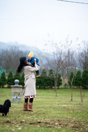 Against a muted background, a woman in a coat holds a toddler high in the air. A small dog stands on the lawn nearby. There is a misty mountain in the background, and a cloudy sky.の写真素材