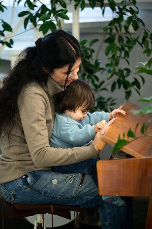 A mother holds her child on her lap as they explore a wooden object. Sunlight streams in, casting a warm glow on them while potted plants surround them, creating a cozy atmosphere.の写真素材