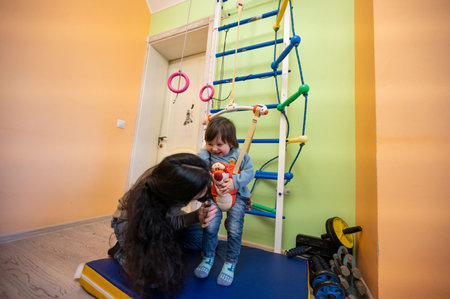 A child smiles brightly while being held by a caregiver in a colorful playroom. Climbing equipment and toys surround them, creating a joyful atmosphere for play and bonding.の写真素材