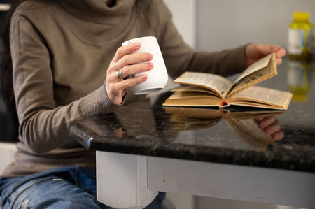 A woman in a comfortable sweater enjoys her morning routine, sipping coffee while immersed in a book at a sleek kitchen table, surrounded by a warm ambiance.の写真素材