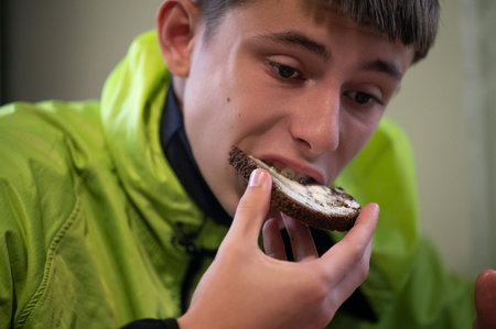 A young individual takes a bite of dark rye bread topped with a unique spread, showcasing a moment of enjoyment in a warm indoor atmosphere amidst a leisurely gathering.の写真素材