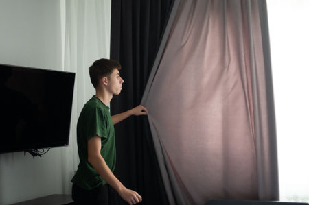 A young man stands near a window, gently pulling back the curtains to let the warm morning sunlight flood into the bright and inviting room.の写真素材