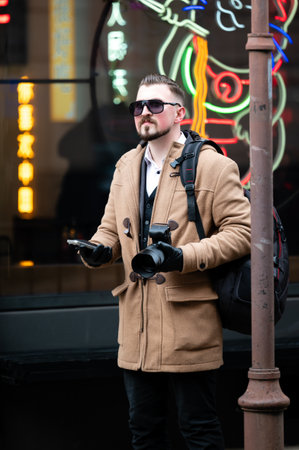 Dressed in a chic beige coat, a young man with sunglasses stands confidently on the sidewalk,...