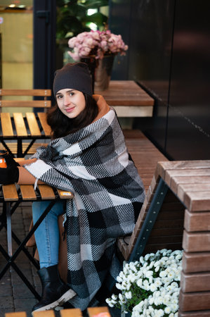 Wrapped in a soft blanket, a young woman enjoys a relaxing afternoon at a charming cafe, surrounded by warm lighting and lovely floral arrangements.の写真素材