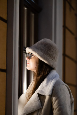 After dusk, a woman stands beside a window, lost in contemplation while wearing a warm coat and a...