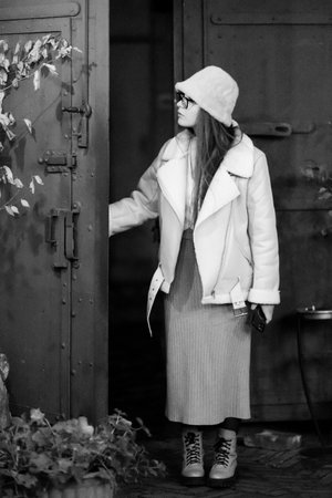 Dressed in a chic winter outfit, the young woman exudes an air of mystery as she leans against a textured metal door, surrounded by greenery in a quaint alleyway.の写真素材