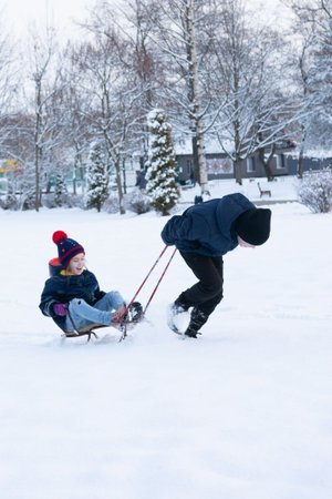 Two children playing in the snow. One child is sitting on a sled while the other pulls them along in...