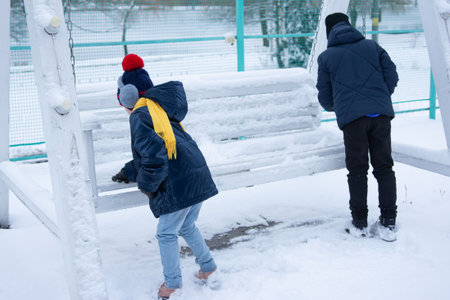 Two children in winter clothing are playing near a snow-covered white swing set. One child examines the snow, the other stands nearby. The scene is outdoors in a snowy environment.の写真素材