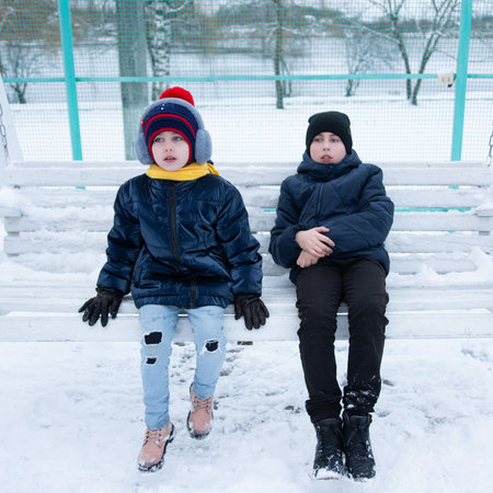 Two children, a boy and a girl, dressed warmly in winter coats and hats, sit side-by-side on a white bench covered in snow. The serene winter scene provides a backdrop to their quiet contemplation.の写真素材