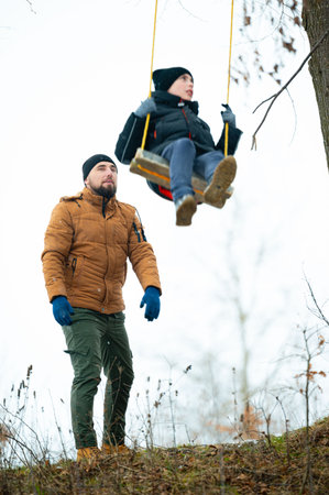 A man in a brown jacket observes his child swinging on a homemade wooden swing hanging from a tree branch in a snowy, wintry setting. The scene is peaceful and heartwarming.の写真素材