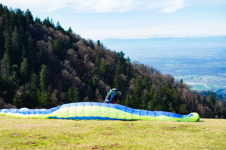 A paragliding enthusiast meticulously prepares their colorful wing on a grassy mound, ready to take...