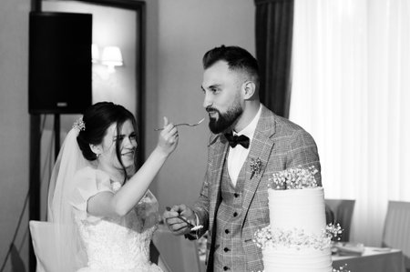 A black and white photo of a bride and groom at their wedding reception. The bride is feeding the groom a piece of wedding cake with a playful smile.の写真素材