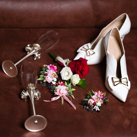 A close-up shot showcasing wedding essentials: a pair of white heels, two ornate champagne flutes, and several delicately crafted floral boutonnieres. The items are arranged on a rich brown surface.の写真素材