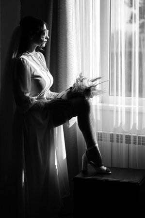A black and white photograph of a bride in a flowing white gown, sitting by a window, holding her wedding bouquet. Sunlight streams through the curtains, illuminating her figure.の写真素材