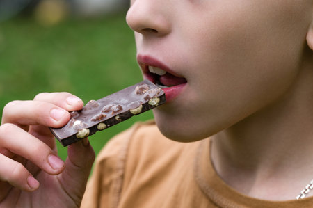 Close-up of a child taking a bite of a chocolate bar studded with nuts. The child is outdoors, with a blurred green background, possibly a park or garden.の写真素材