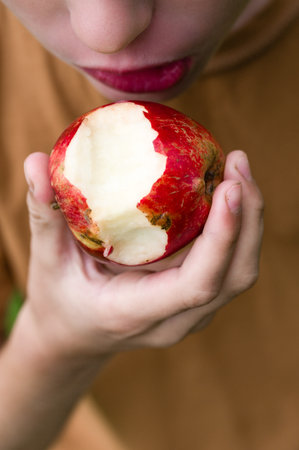 Close-up of a person holding and eating a partially eaten red fruit. The focus is on the fruit and the person's hands.の写真素材