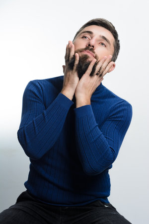 A bearded man in a blue sweater and dark pants sits against a white backdrop, gently stroking his beard with both hands, his gaze directed upwards in contemplation.の写真素材