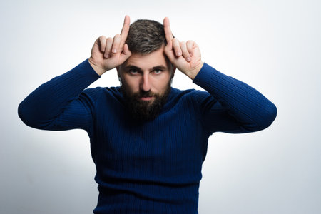 A bearded man in a blue sweater gestures with his fingers, creating 'horns' above his head against a plain background. He looks directly at the camera with a serious expression.の写真素材
