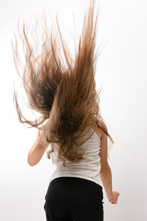 A young girl, seen from behind, with long brown hair dynamically tossed over her shoulders. She wears a white tank top and black pants against a white background.の写真素材