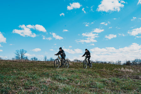 Two people ride mountain bikes across a gently sloping grassy hill. The sky is a vibrant blue with fluffy white clouds. A peaceful, outdoor activity.の写真素材