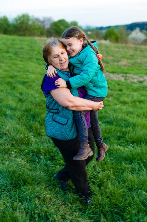 A loving grandmother playfully lifts her granddaughter in a grassy field. The scene exudes warmth...