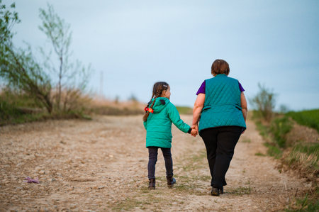 A tender image of a child and adult walking hand-in-hand along a dirt road. The setting is rural, with grassy fields visible in the distance. They walk away from the camera.の写真素材