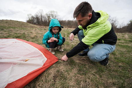A father guides his daughter as they work together to pitch a tent on a grassy field, surrounded by nature and overcast skies, creating a bonding experience.の写真素材