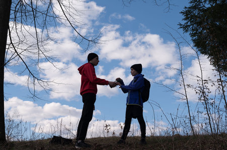 Silhouetted figures of two young people exchanging an item in a natural setting. The sky is bright...