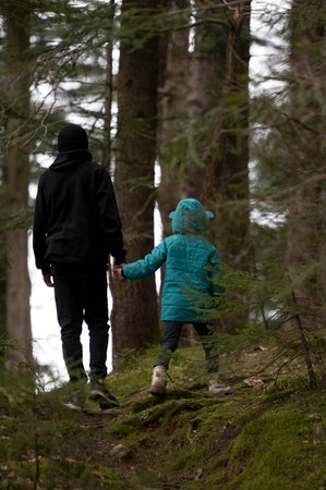 A child, adorned in a teal jacket, holds hands with an adult as they stroll through a lush green forest surrounded by towering trees and soft moss underfoot.の写真素材