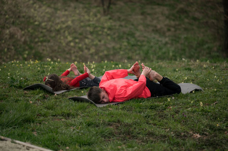 Two children lie side by side on mats in a lush green park, engaging in playful stretches while surrounded by nature, embracing the joy of outdoor yoga practice.の写真素材