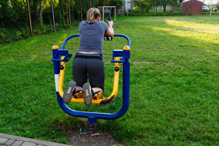 A woman uses an outdoor exercise machine in a park setting. The machine is blue and yellow, and she is wearing athletic clothing. The grass is green and well-maintained.の写真素材