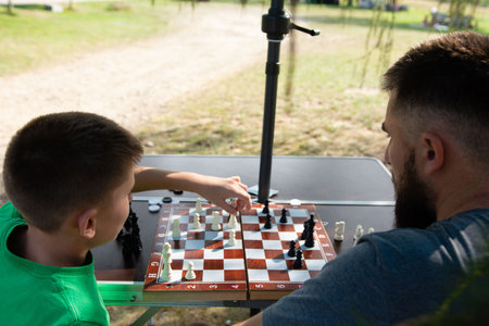 Father and son engage in a friendly game of chess on a portable board set up outside. The relaxed setting suggests leisure time spent together.の写真素材