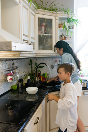 A woman and boy collaborate on washing dishes in a modern kitchen with white cabinets and dark countertops. They work together, fostering a sense of teamwork and family.の写真素材