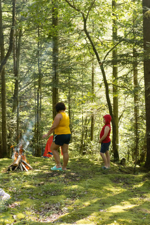 A woman and child pause by a small campfire in a lush green forest. Sunlight filters through the trees, creating a serene atmosphere. A tent is partially visible in the background.の写真素材