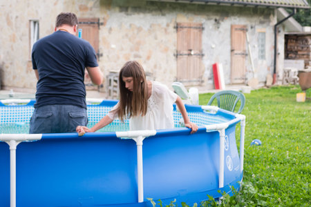 A young girl carefully enters a round, above-ground swimming pool in a grassy backyard. An adult is...