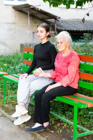 A young woman and her elderly companion sit side by side on a vibrant green bench, sharing a heartfelt moment in a tranquil park during a sunny afternoon.の写真素材