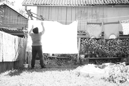 In a black and white composition, an individual hangs a large white sheet on a clothesline in a yard. Behind them, a woodpile and a shed add to the rustic, domestic setting.の写真素材
