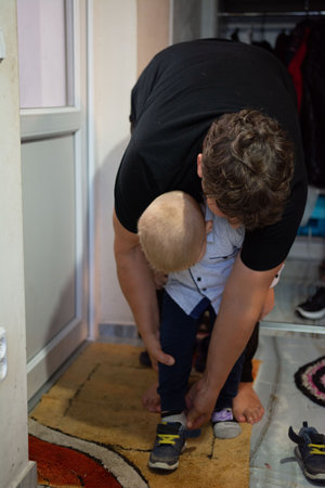 This intimate scene shows a caring parent assisting a young child with putting on their shoes in an entryway. The focus is on the connection and tender moment shared during preparation.の写真素材