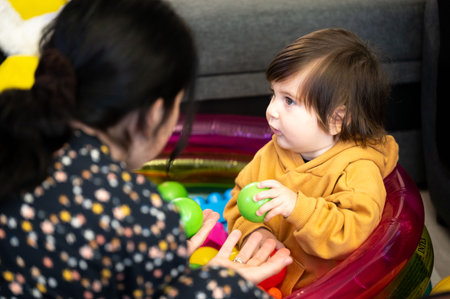 Baby in yellow hoodie sits in colorful chair, playing with caregiver in toy-filled room. Afternoon.