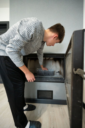 A young man in cozy attire bends over to clean out ashes from a contemporary fireplace, ensuring a tidy living space in the middle of a bright, stylish room.の写真素材