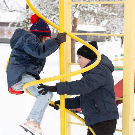 Two children, a boy and a girl, bundled in winter clothes, enjoy a snowy day playing on a yellow spiral climbing frame. They are actively engaged, one assisting the other.の写真素材