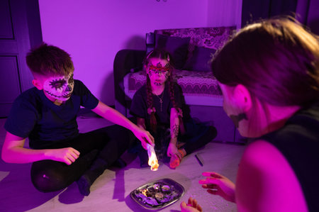 Three children, faces painted like skulls, sit on a floor illuminated in purple light. They're...