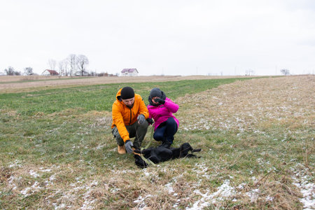 A man in an orange jacket and a woman in a pink coat interact with their black dog in a snowy field,...
