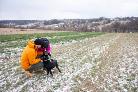Amidst a chilly winter day, two friends interact warmly with a playful black dog on a grassy patch...