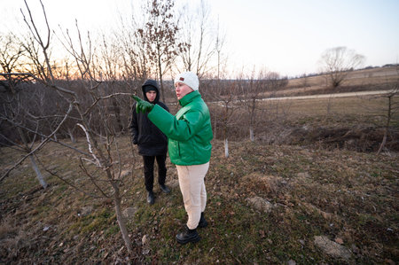 Two individuals engage in conversation about tree care in a serene rural area as daylight fades. The...
