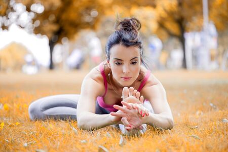 Young woman practicing yoga in the park on the green grass with fallen leaves in autumn with blurred background. Shallow depth of fieldの写真素材