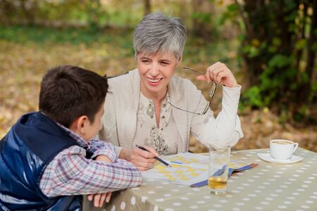 Older woman with gray hair sitting with grandson in the park reading a book, drinking coffee and talkingの写真素材
