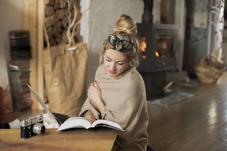 Young beautiful woman in the living room sits on the floor and communicates with friends over the computer, covered with a blanket for the cold in the back of the fireplaceの写真素材